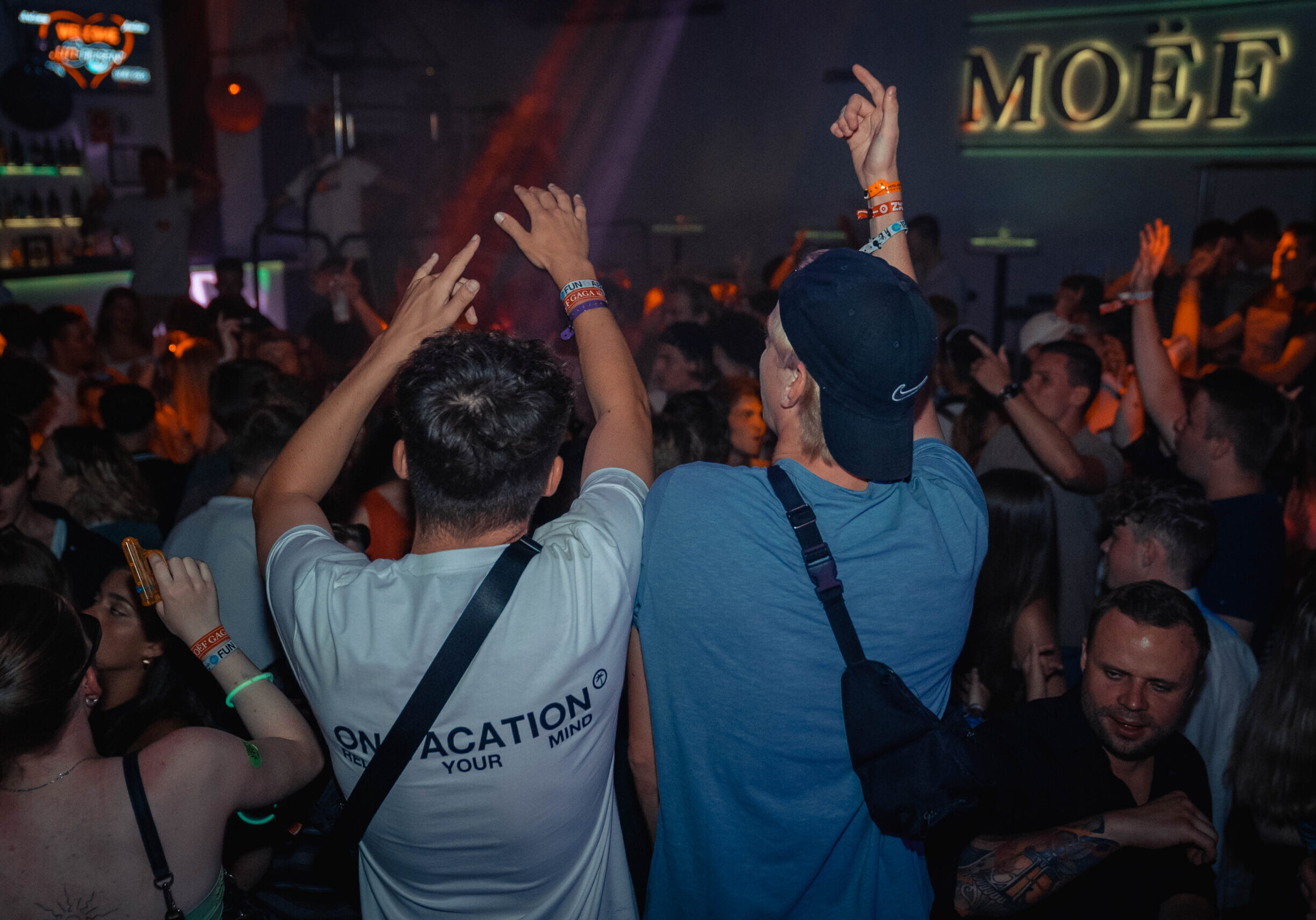 Zwei junge Männer stehen von hinten gesehen in einer Menschenmenge, die in einem Nachtclub tanzt und feiert, mit erhobenen Händen zur Musik – die pure Energie einer Jugendreise-Party.