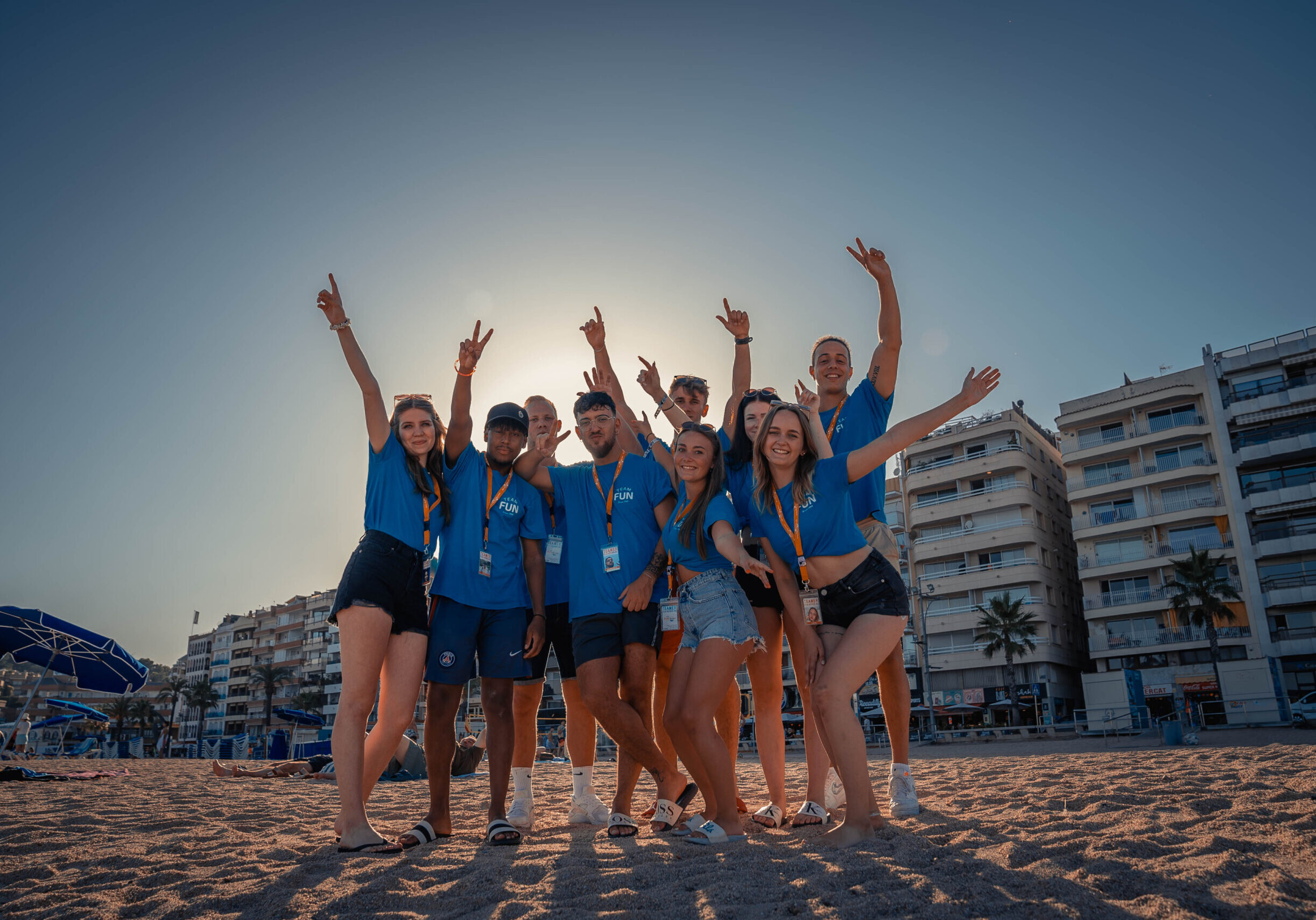 Eine fröhliche Gruppe von neun jungen Menschen in blauen FUN Reisen T-Shirts und Shorts steht am Strand vor einer Reihe von Gebäuden, lacht und jubelt mit erhobenen Armen, während die Sonne im Hintergrund strahlt