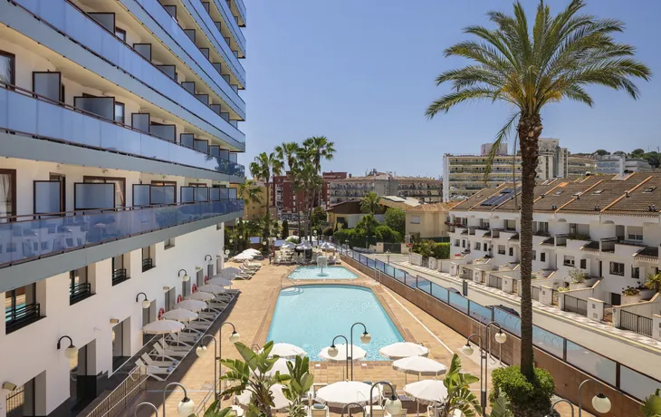 Außenansicht eines Hotels mit Poolbereich, Palmen und umliegenden Gebäuden unter blauem Himmel.