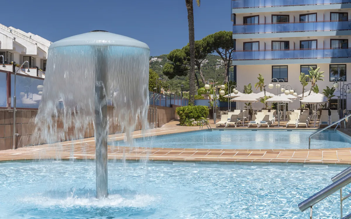 Außenansicht eines Hotelpools mit Pilz-Wasserfall, Liegen und Hotelgebäude unter blauem Himmel.